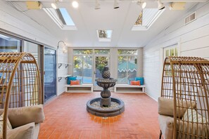 A sunlit room with a central fountain, seating areas with cushions, and a view through large windows. - Riverwalk Villa Game Room Grill Heated Pool (San Antonio)