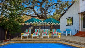 A swimming pool with a tiled edge, surrounded by outdoor seating with colorful cushions and a large tree providing shade. - Riverwalk Villa Game Room Grill Heated Pool (San Antonio)