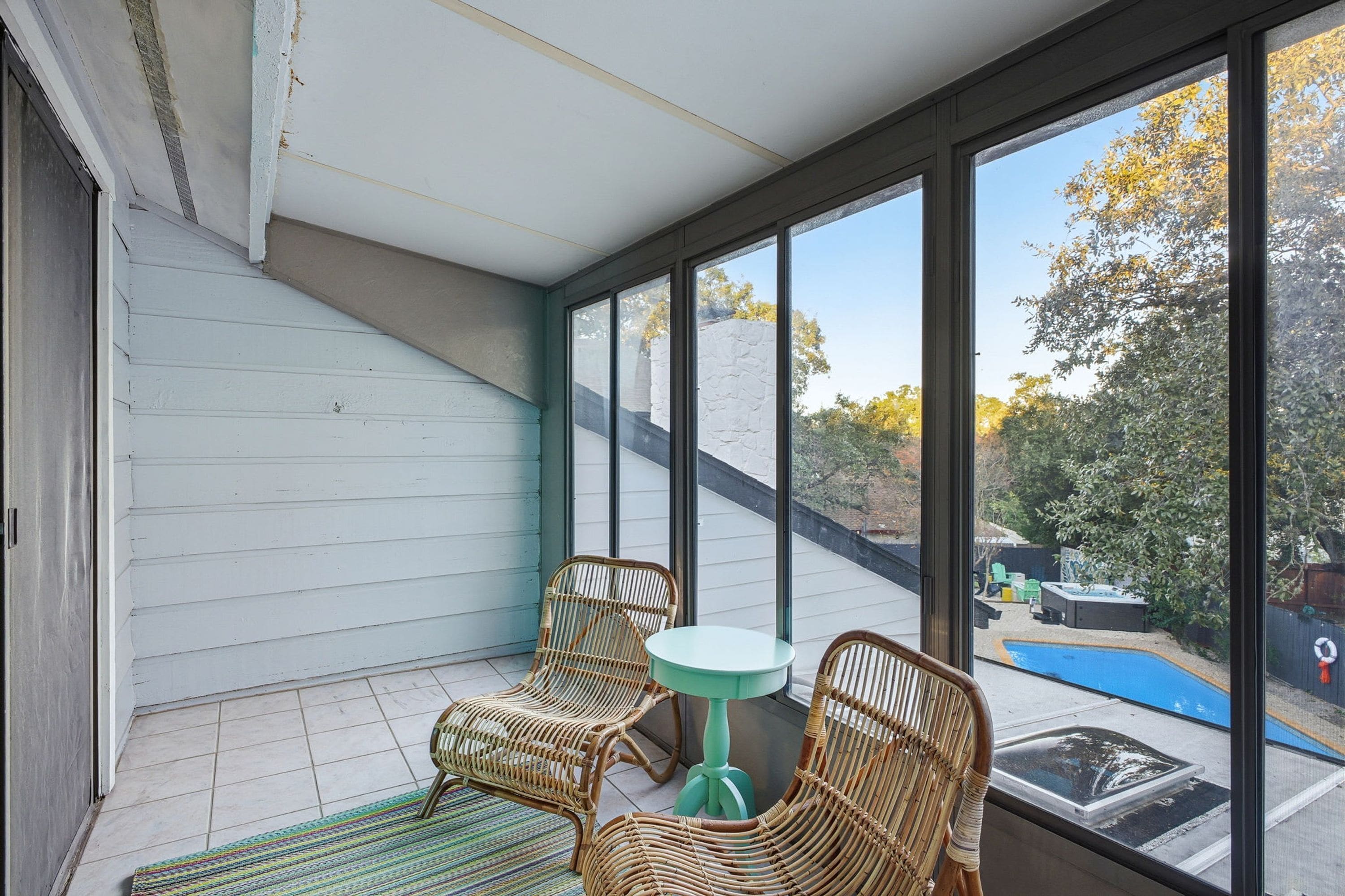 A sunroom with two wicker chairs and a small table, large windows, and a view of a backyard with a pool.