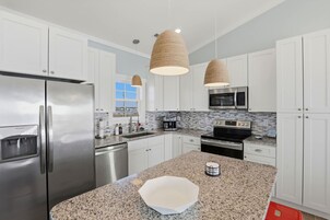 Cocina moderna con gabinetes blancos, electrodomésticos de acero inoxidable, encimera de granito y dos lámparas colgantes tejidas.