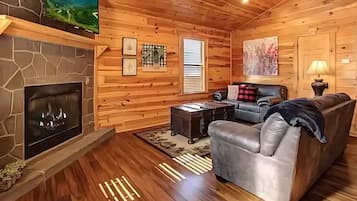 A cozy cabin interior with a stone fireplace, wooden walls, a leather sofa, and a television mounted on the wall.