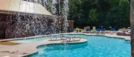 A pool with a waterfall feature, surrounded by lounge chairs and a wooded area.