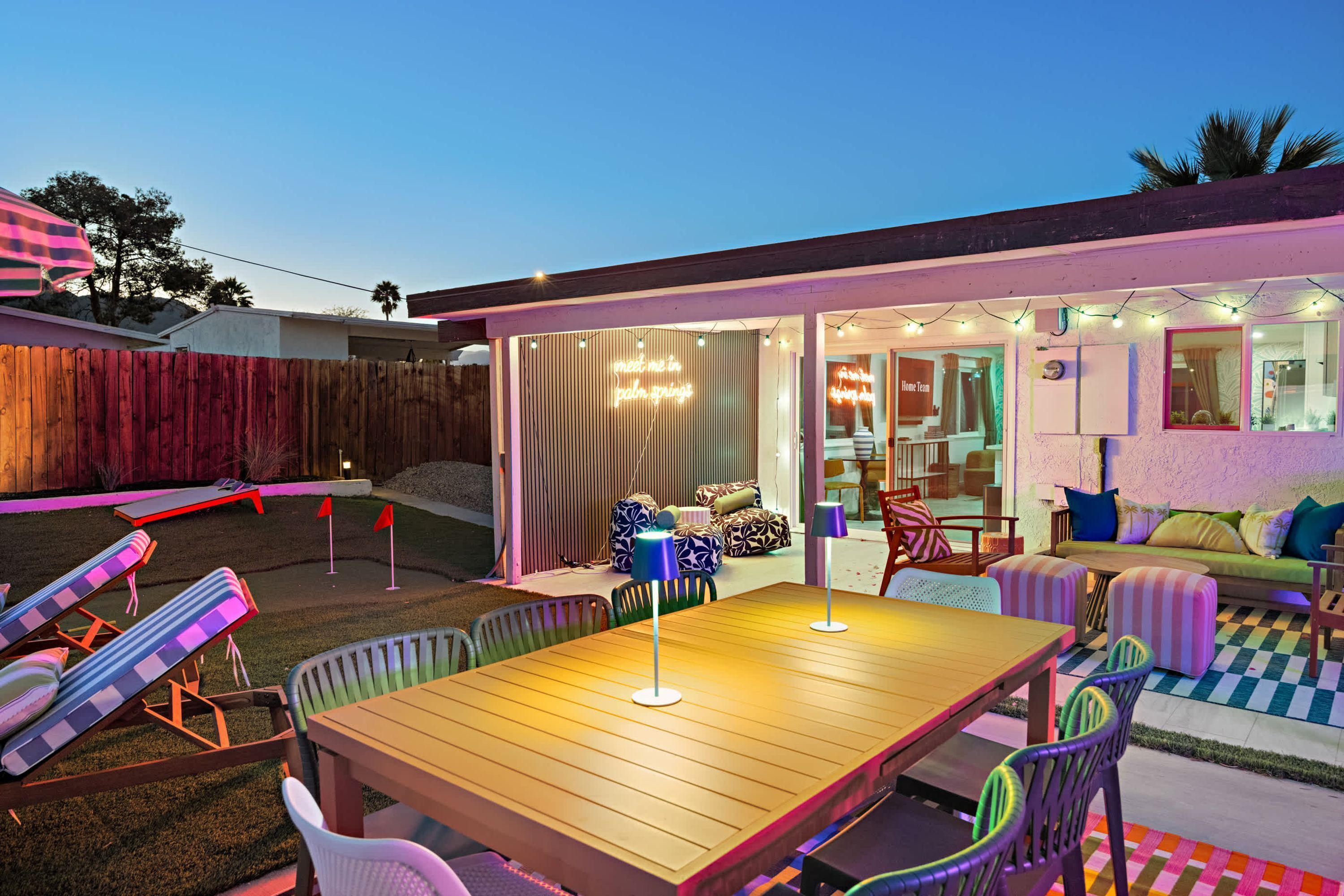 Un patio al aire libre con una mesa y sillas de madera, una zona de estar cubierta con cojines y vistas hacia la casa.