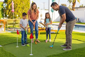 Eine Familie spielt Minigolf im Freien, im Hintergrund ist ein Schwimmbad zu sehen.