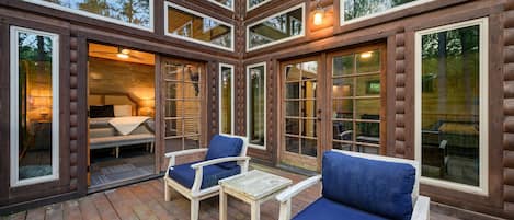 A log cabin with a deck, two chairs, a table, and a bed with a view of the forest.