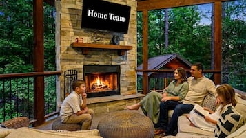 A stone fireplace with a television labeled 'Home Team' above it, surrounded by a wooden deck and outdoor seating.
