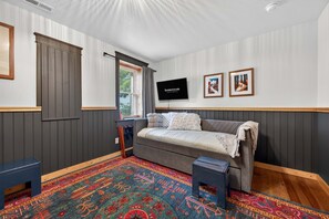 Ein Zimmer mit einem Bett, einem Fernseher, einem Fenster mit Jalousien und einem farbenfrohen Teppich.