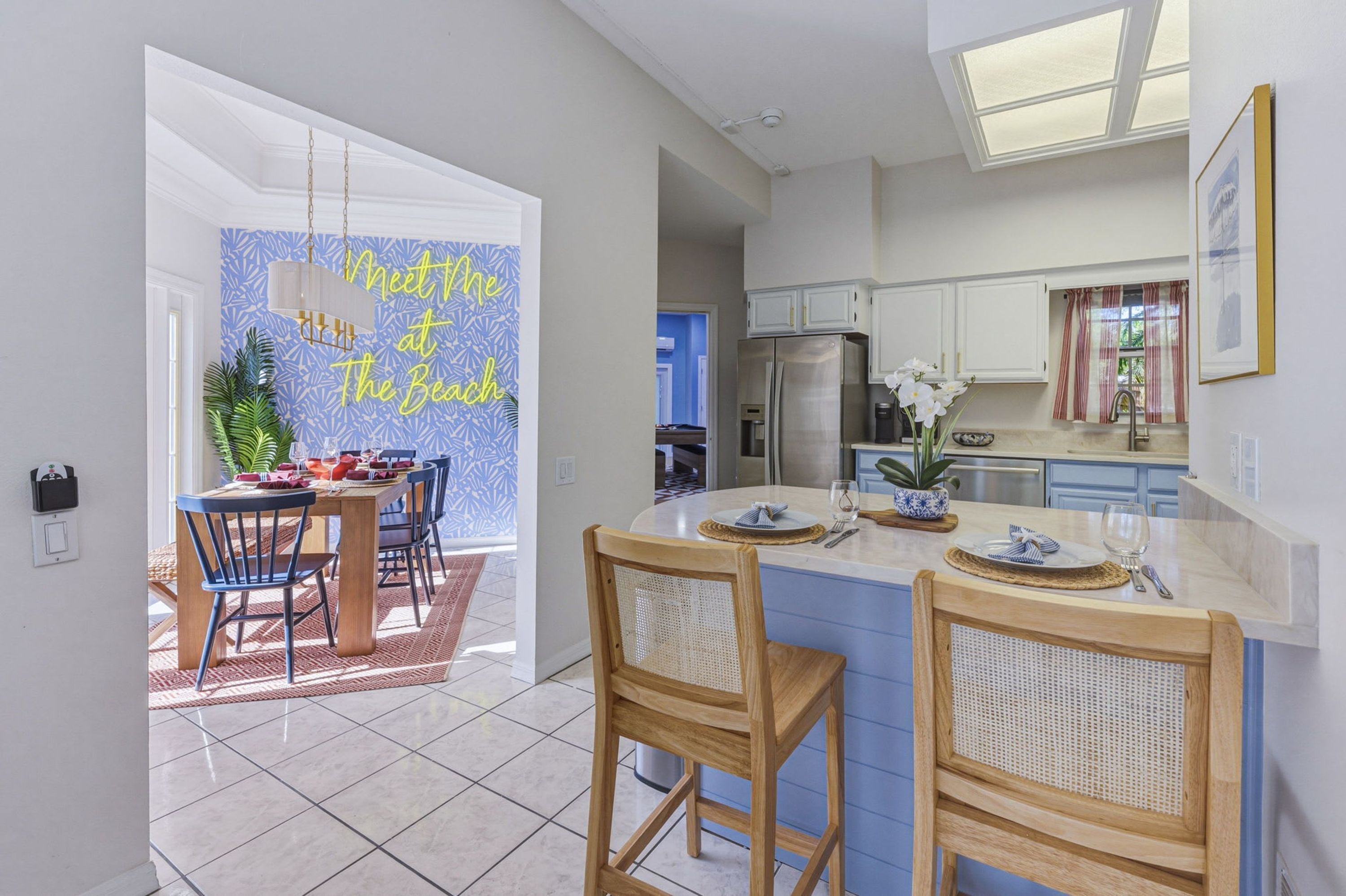 A modern kitchen with a central island, white cabinets, and a dining area with a blue wall and a 'Meet Me at The Beach' sign.