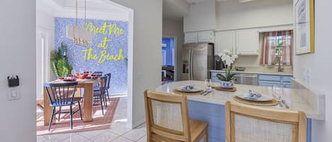 Una cocina moderna con una isla central, armarios blancos y una zona de comedor con una pared azul y un letrero que dice 'Meet Me at The Beach'.