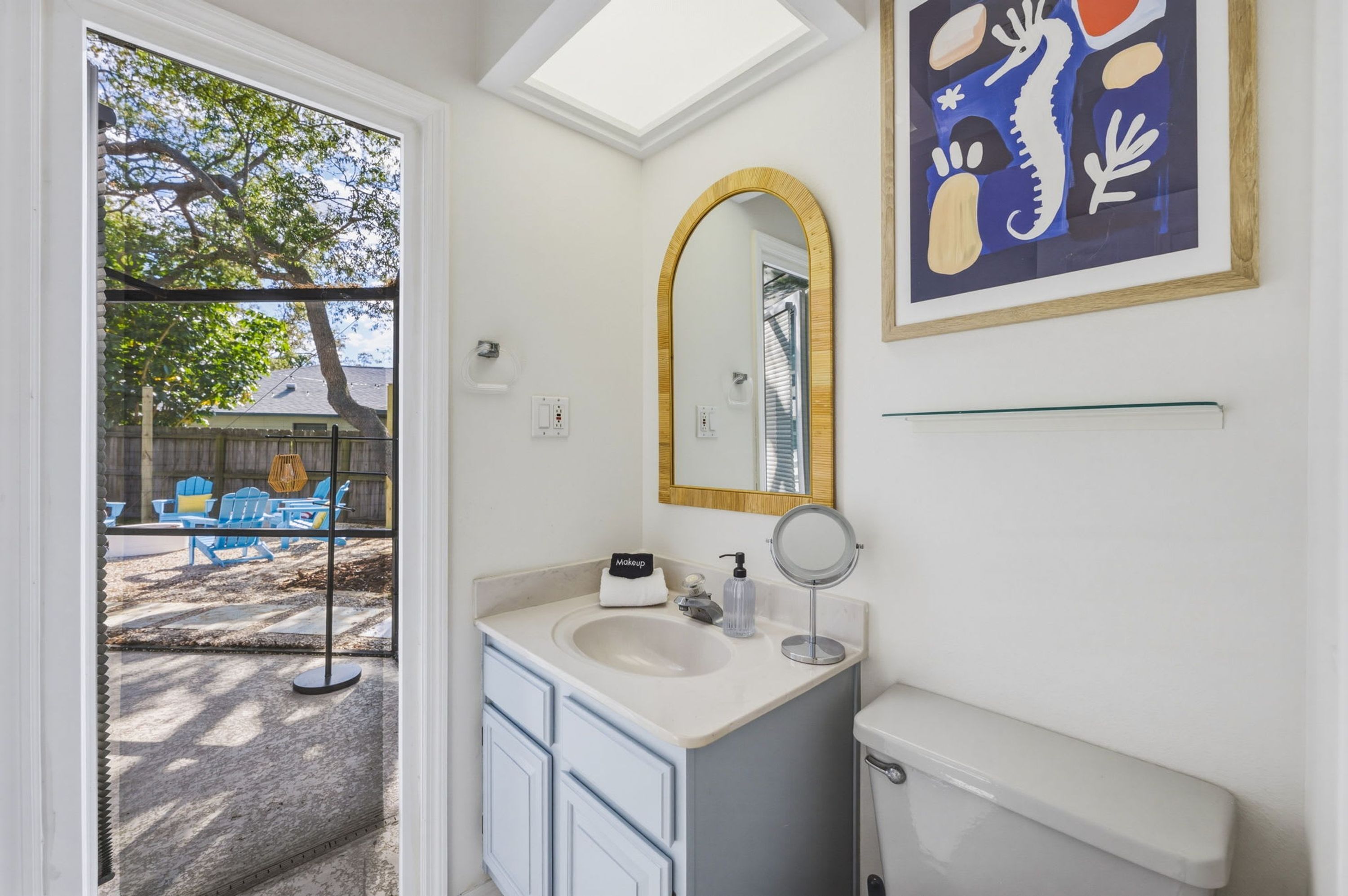 Une salle de bain avec un lavabo blanc, un miroir et des toilettes. Il y a une fenêtre donnant sur du mobilier d’extérieur et des arbres.