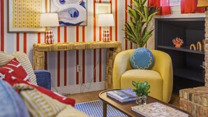 Ein Wohnzimmer mit Streifenmuster-Tapete, einem gelben Sessel, einem blauen Kissen, einem Couchtisch mit einem Buch und einem Fernseher an einer Ziegelwand.