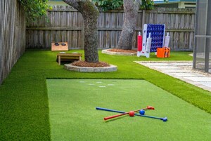Un patio trasero con césped cuidado, un banco de madera, un árbol y un área de juegos pequeña con una pelota y un juego de croquet.