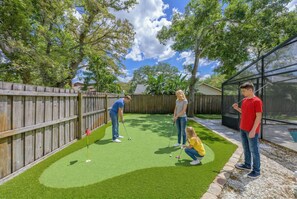 Ein Putting Green im Freien mit Holzzaun, Glasumrandung und Bäumen im Hintergrund.