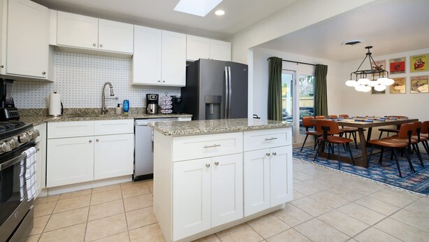 A modern kitchen with white cabinetry, a central island, and a dining area with a table and chairs. - Starcrest 4 Br Haven Pool Hot Tub Putt Putt Arcade (San Antonio)