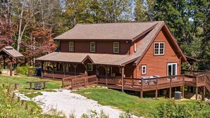 A two-story log cabin with a porch and a deck, surrounded by trees and a gravel path. - Luxury 8BR Mountain Lodge Hot Tub Fire Pit (Byrdstown)