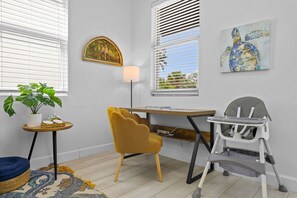 Ein Zimmer mit Schreibtisch aus Holz, einem gelben Stuhl, einem Hochstuhl, einer Lampe, einem Gemälde und einer Topfpflanze.