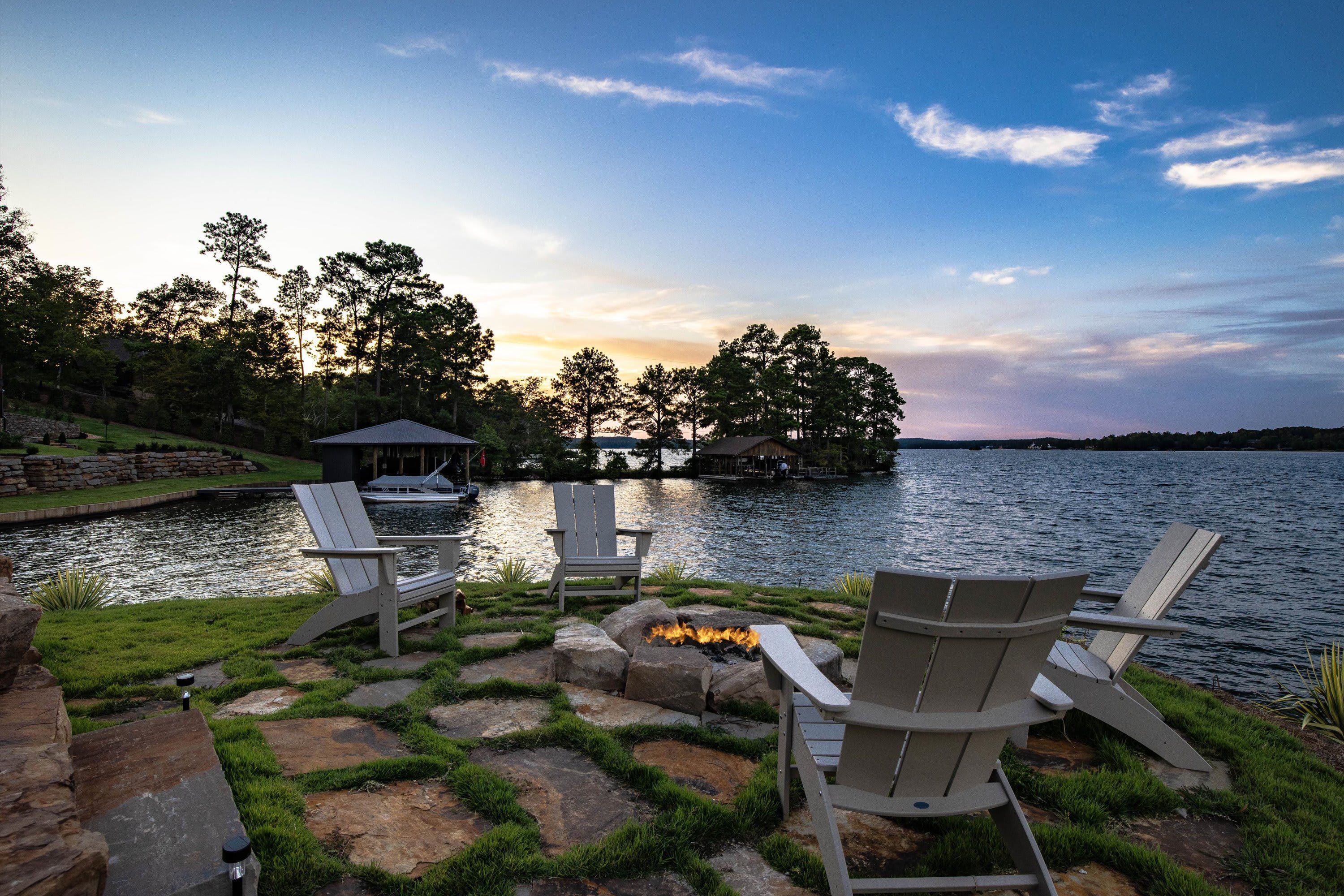 A lakeside deck with Adirondack chairs, a fire pit, and a dock extending into the water.