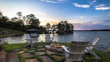 A lakeside deck with Adirondack chairs, a fire pit, and a dock extending into the water.