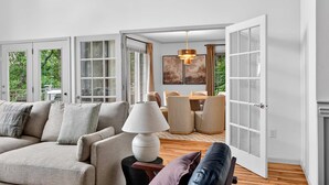 A spacious living room with a sofa, a dining table, and a coffee table. There is a large window with a view of greenery outside. - Luxury Mtn Retreat Hot Tub Sleeps 18 Game Room (Asheville)