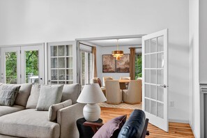 A spacious living room with a sofa, a dining table, and a coffee table. There is a large window with a view of greenery outside. - Luxury Mtn Retreat Hot Tub Sleeps 18 Game Room (Asheville)