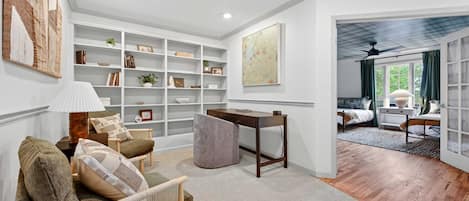 Uma sala de estar espaçosa com uma grande estante de livros, um confortável espaço de estar com um sofá e poltronas, e vista para um quarto com cama e janela.