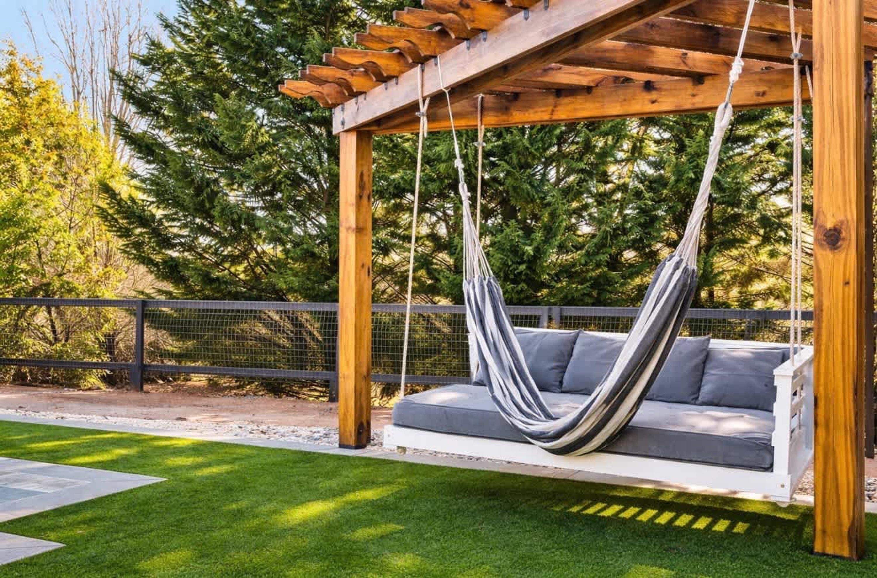 A hammock swing attached to a wooden pergola, with a grey cushioned bench below.