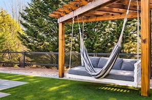 Una hamaca colgada de una pérgola de madera, con un banco acolchado gris debajo.