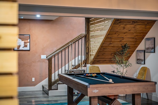 Una mesa de billar en una habitación con una escalera de madera y vistas al exterior.
