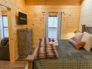 A wooden cabin bedroom with a bed, a dresser, a TV, and a window with curtains. - Smoky Mtn Retreat Sauna Hot Tub Game Room (Pigeon Forge)