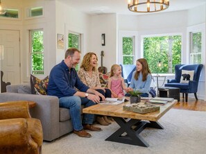 A family sitting on a couch in a well-lit room with large windows, a wooden coffee table, and a blue armchair. - Mountain Views Hot Tub Firepit Game Room (Asheville)