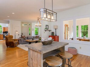 A spacious living area with a wooden coffee table, a leather armchair, and a sofa. There is a large window providing natural light and a view of greenery outside. - Mountain Views Hot Tub Firepit Game Room (Asheville)