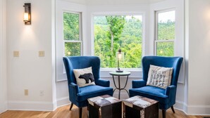 A room with two blue armchairs, a small round table, and a view of a forest through large windows. - Mountain Views Hot Tub Firepit Game Room (Asheville)