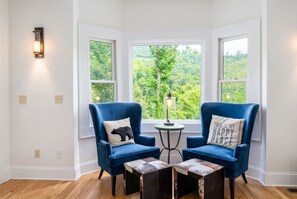 A room with two blue armchairs, a small round table, and a view of a forest through large windows. - Mountain Views Hot Tub Firepit Game Room (Asheville)