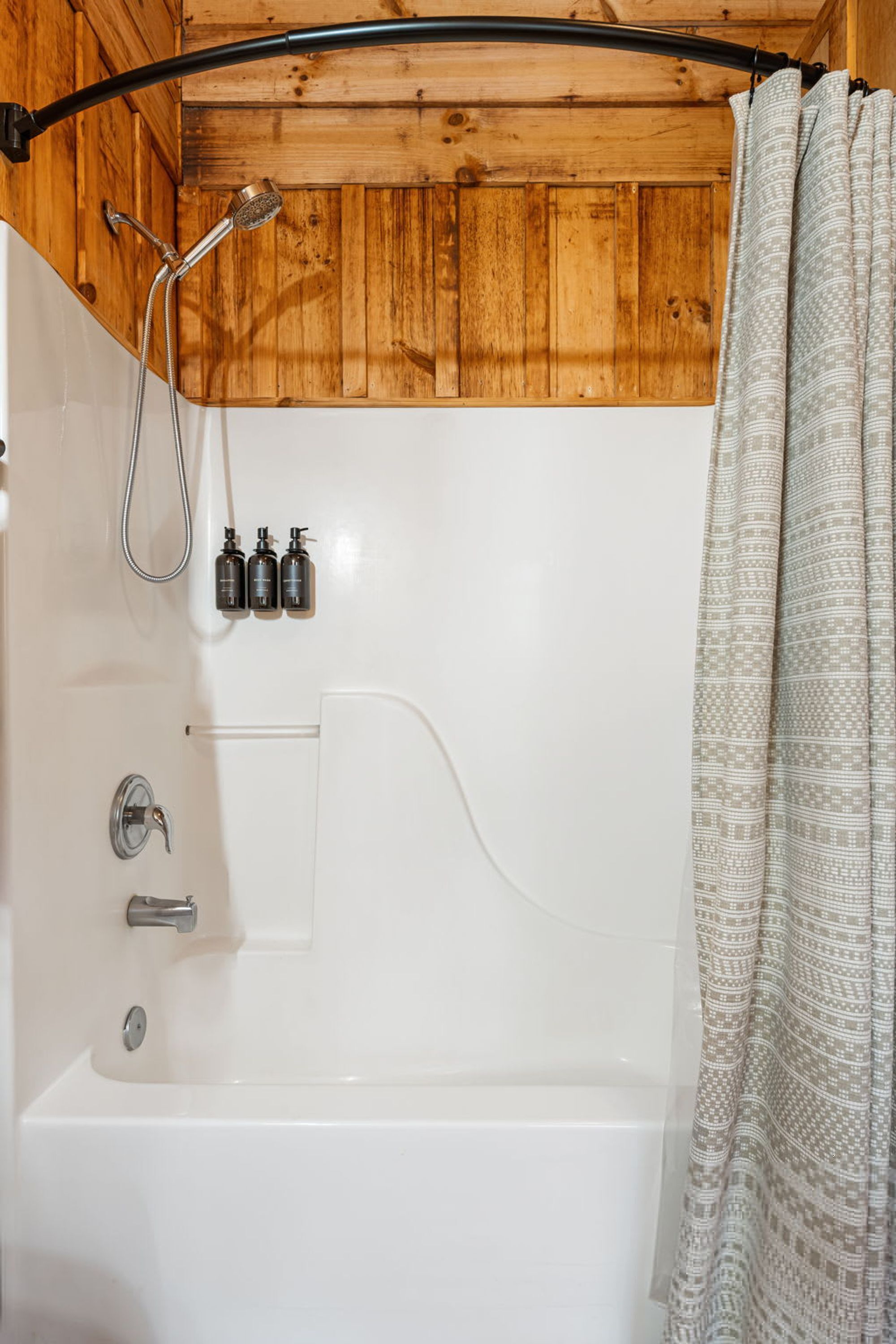 A bathroom with a wooden ceiling, a bathtub, a showerhead, and three bottles of shampoo.