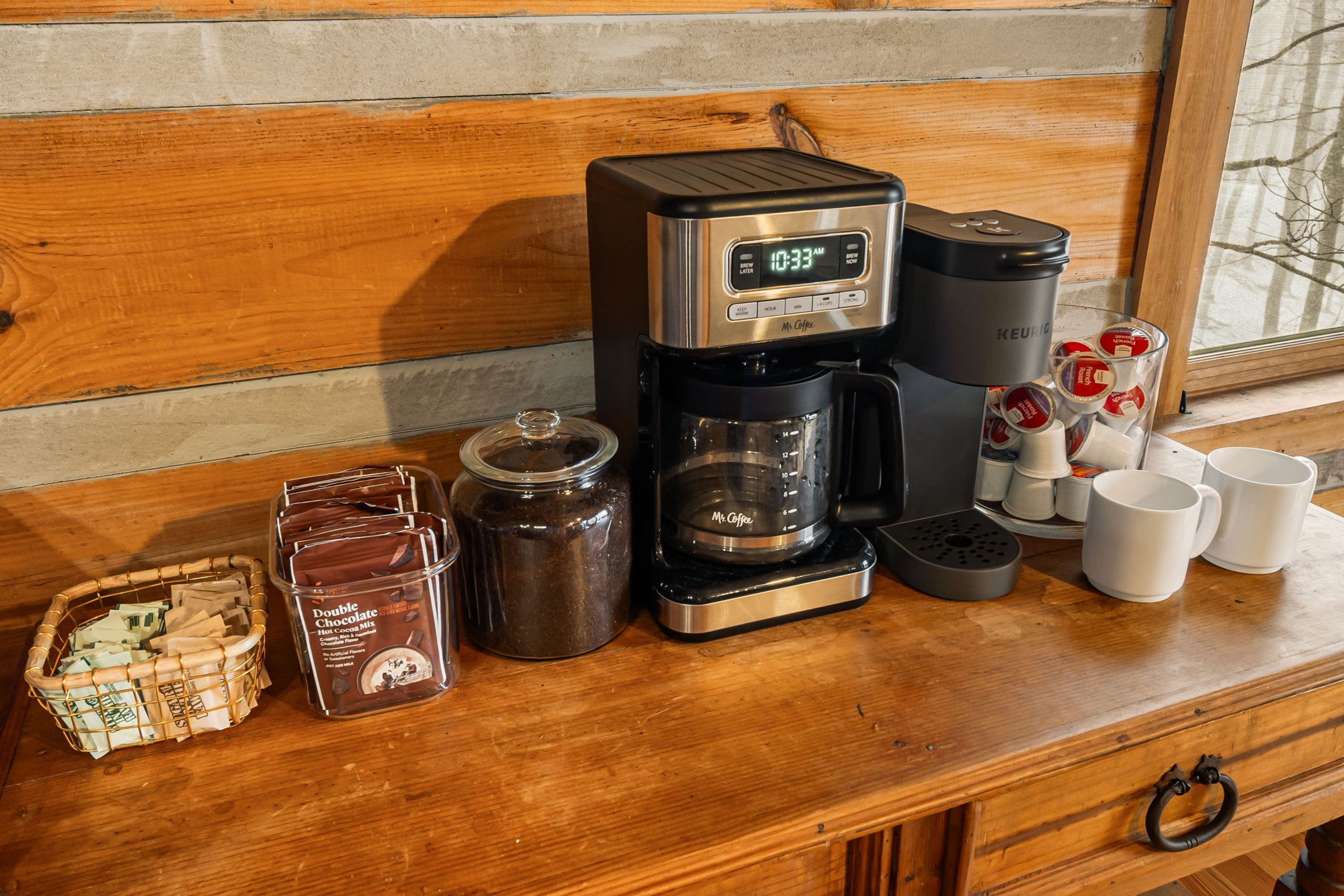 A coffee maker with a digital display, a Mr. Coffee brand coffee pot, a jar of Double Chocolate Hot Cocoa Mix, a basket of coffee pods, and several white mugs on a wooden surface.