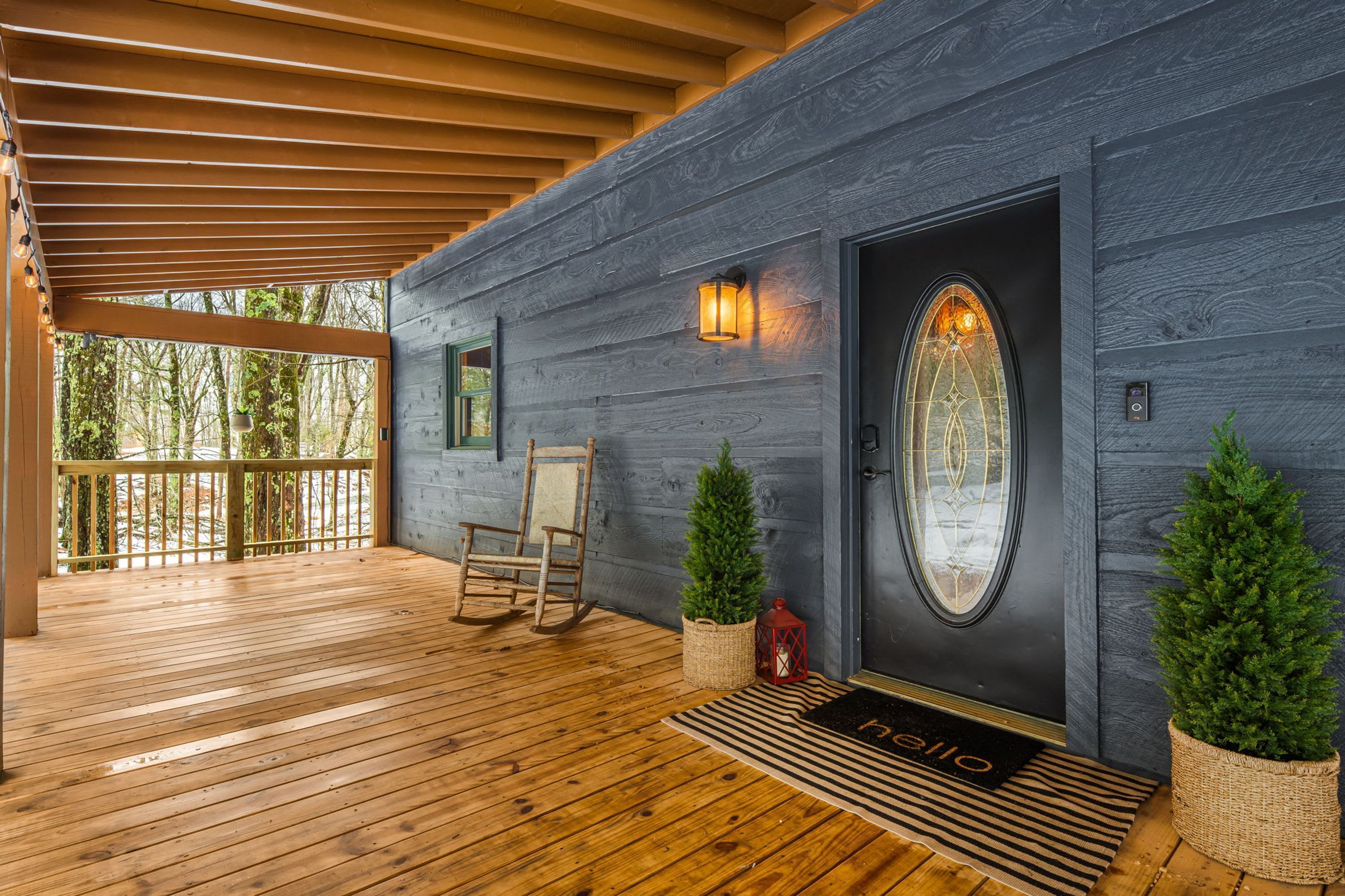 Eine Holzterrasse mit einer dunkelgrauen Tür, einem Schaukelstuhl, zwei Blumentöpfen und einer Fußmatte.