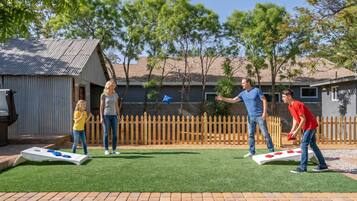 Una familia jugando un juego en el césped artificial de un patio trasero, con una cerca de madera y un cobertizo de metal.