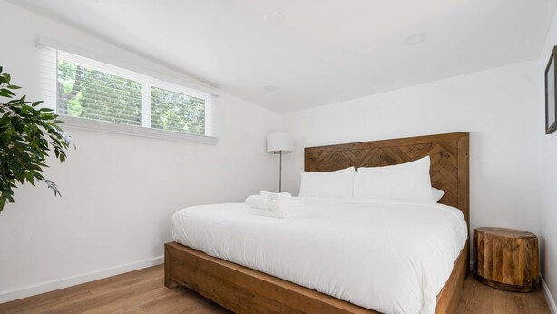 Een slaapkamer met een houten bedframe, wit beddengoed, een bedlampje en een raam met jaloezieën.