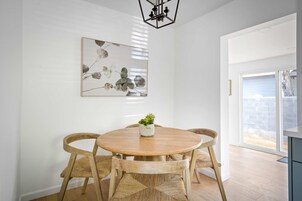 Un comedor con una mesa redonda de madera, cuatro sillas tejidas, una planta en maceta y una obra de arte abstracta colgada en la pared.