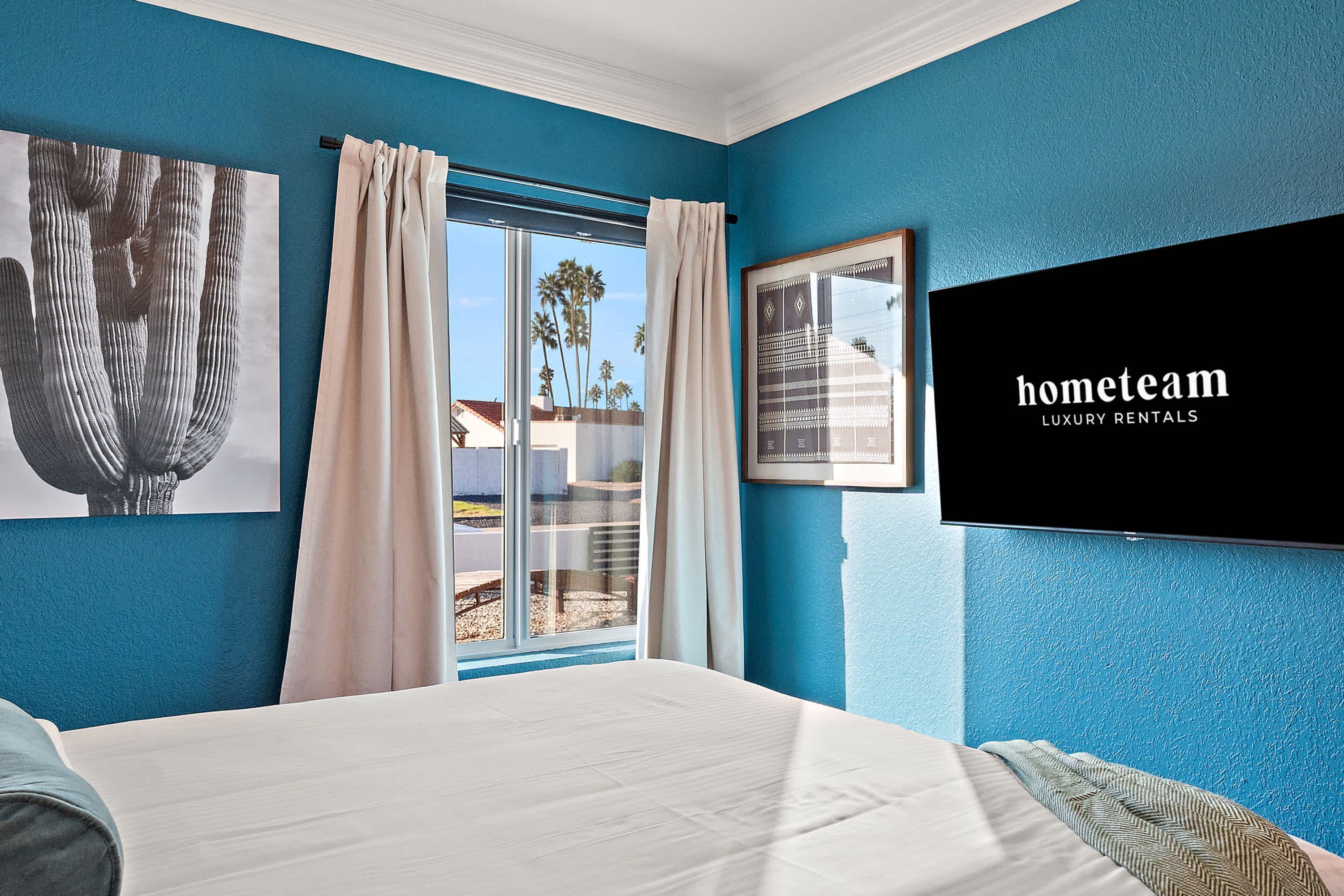 Un dormitorio con una cama grande, una ventana con cortinas y un televisor de pared que muestra 'hometeam LUXURY RENTALS'.