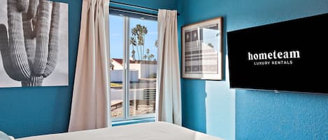 Un dormitorio con una cama grande, una ventana con cortinas y un televisor de pared que muestra 'hometeam LUXURY RENTALS'.