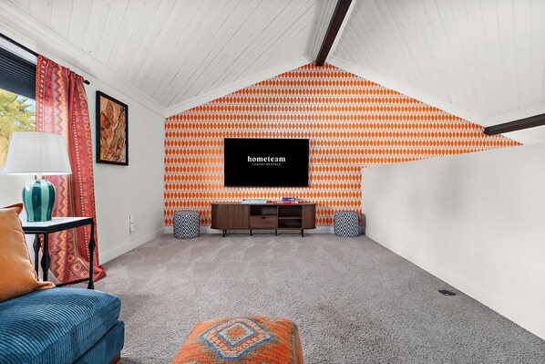 A room with a sloped ceiling, a flat-screen TV mounted on the wall, a wooden cabinet, and patterned cushions. - The Desert Daydream Heated Pool Water Slide (Scottsdale)