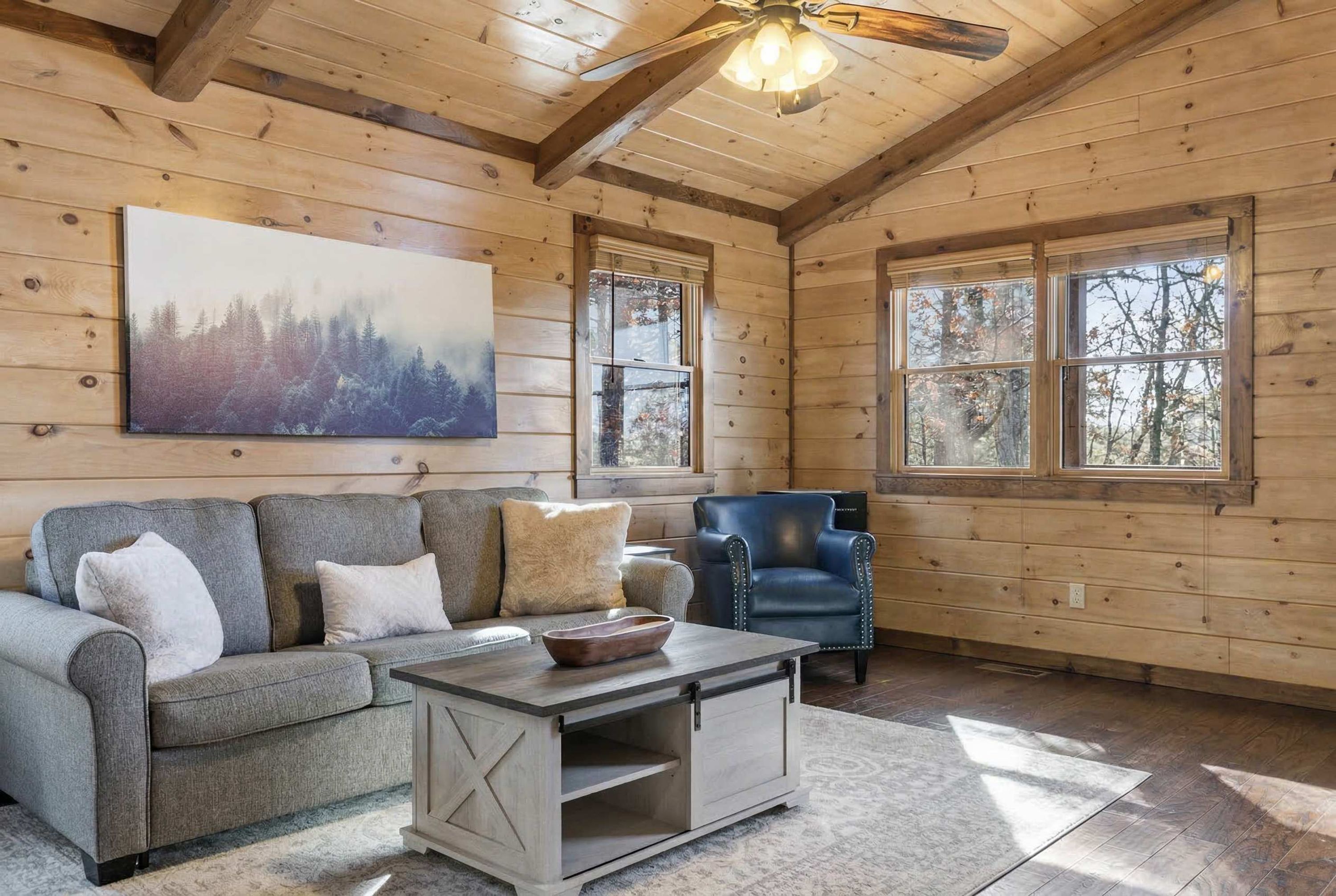 A cozy cabin interior with a wooden ceiling, a large painting of a forest, a gray sofa, a blue armchair, and a wooden coffee table.