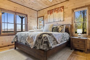 Un dormitorio de cabaña de madera con una cama grande, una mesita de noche y un ventanal con vistas a árboles.