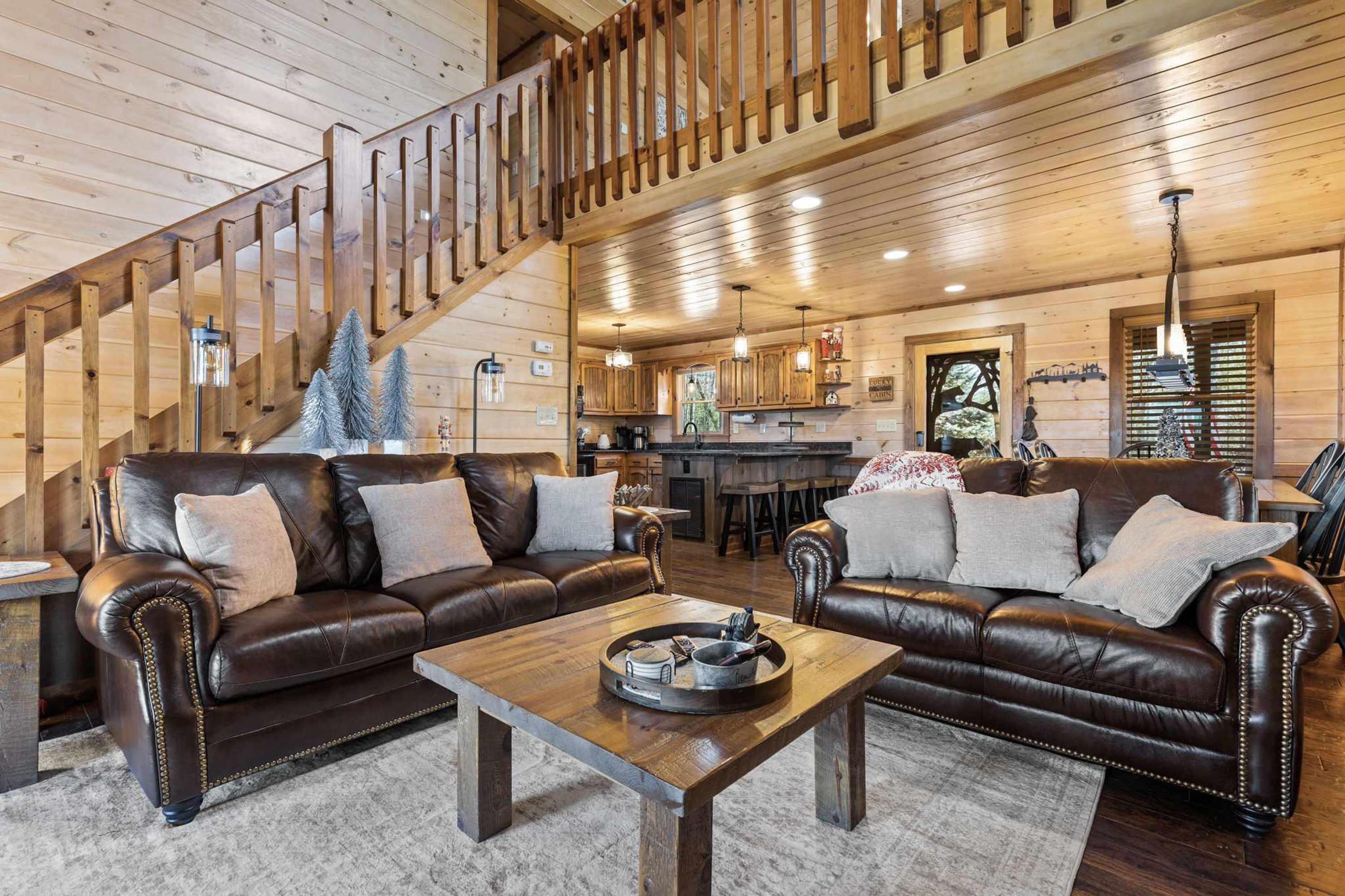 A cozy cabin interior with a wooden staircase, leather sofas, a dining area, and a kitchen.