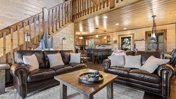 A cozy cabin interior with a wooden staircase, leather sofas, a dining area, and a kitchen.