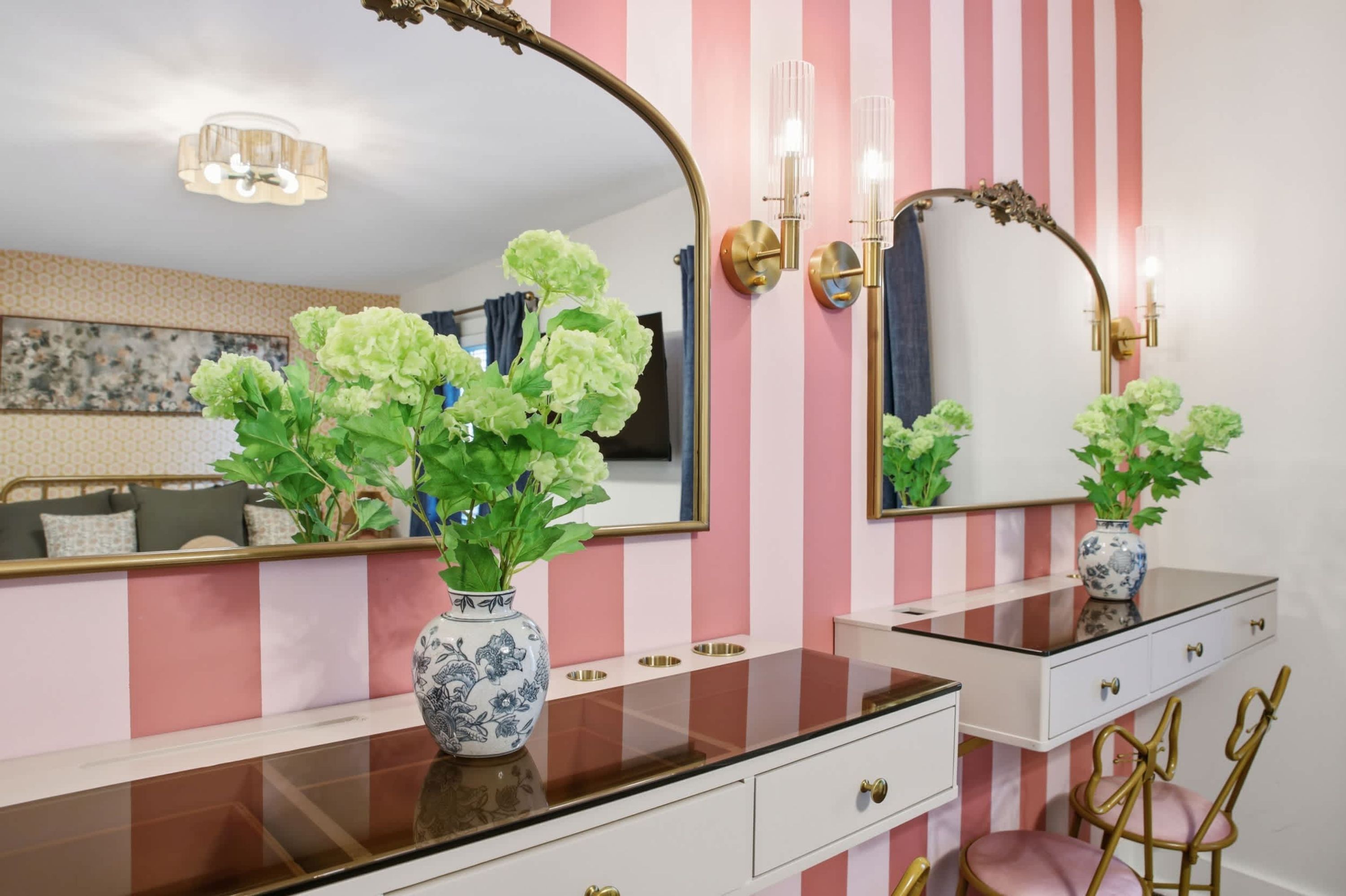A bathroom with a striped wall, a vanity with a mirror, and a vase with green flowers.