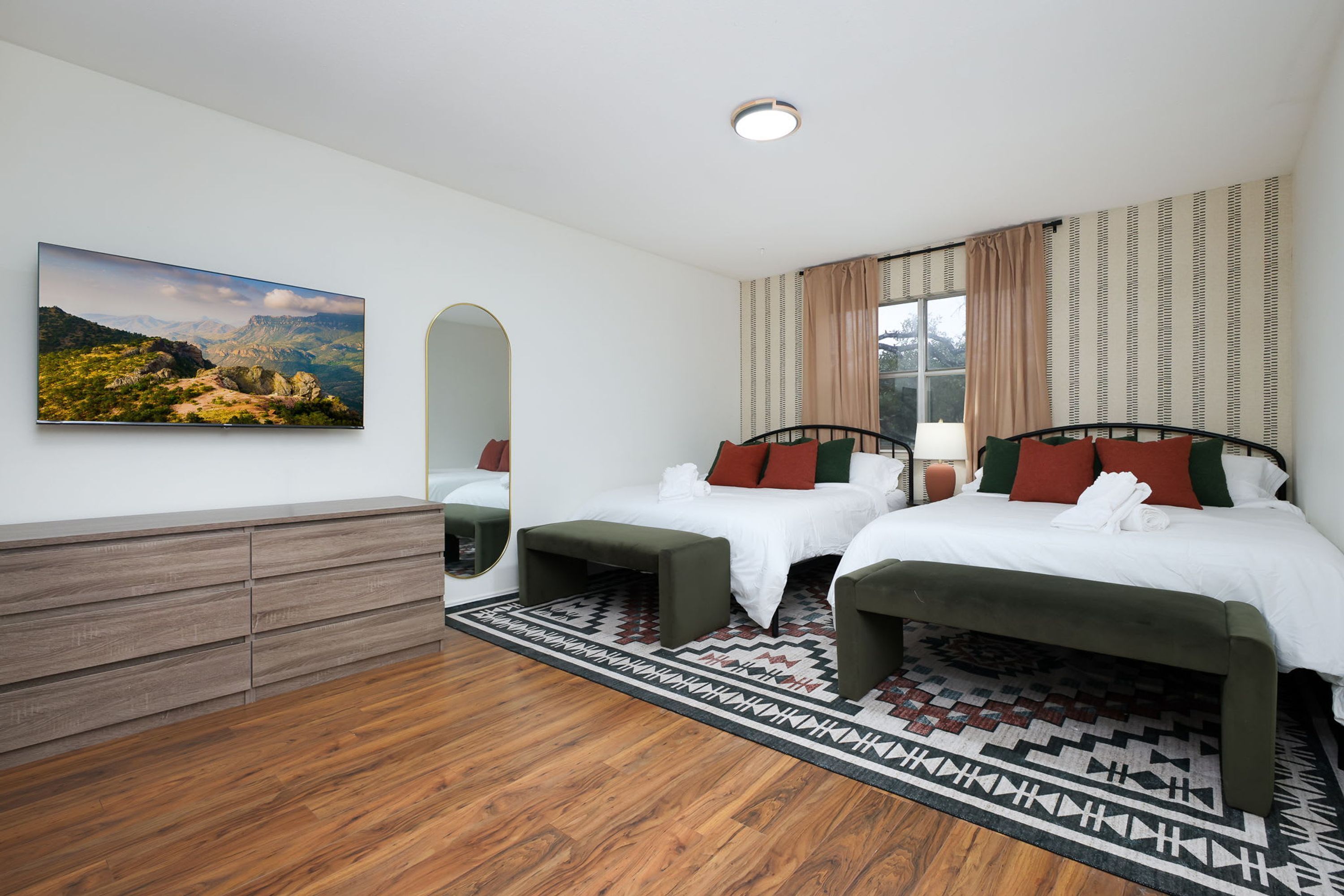 A hotel room with two beds, a wooden dresser, a large mirror, a painting on the wall, and a patterned rug.