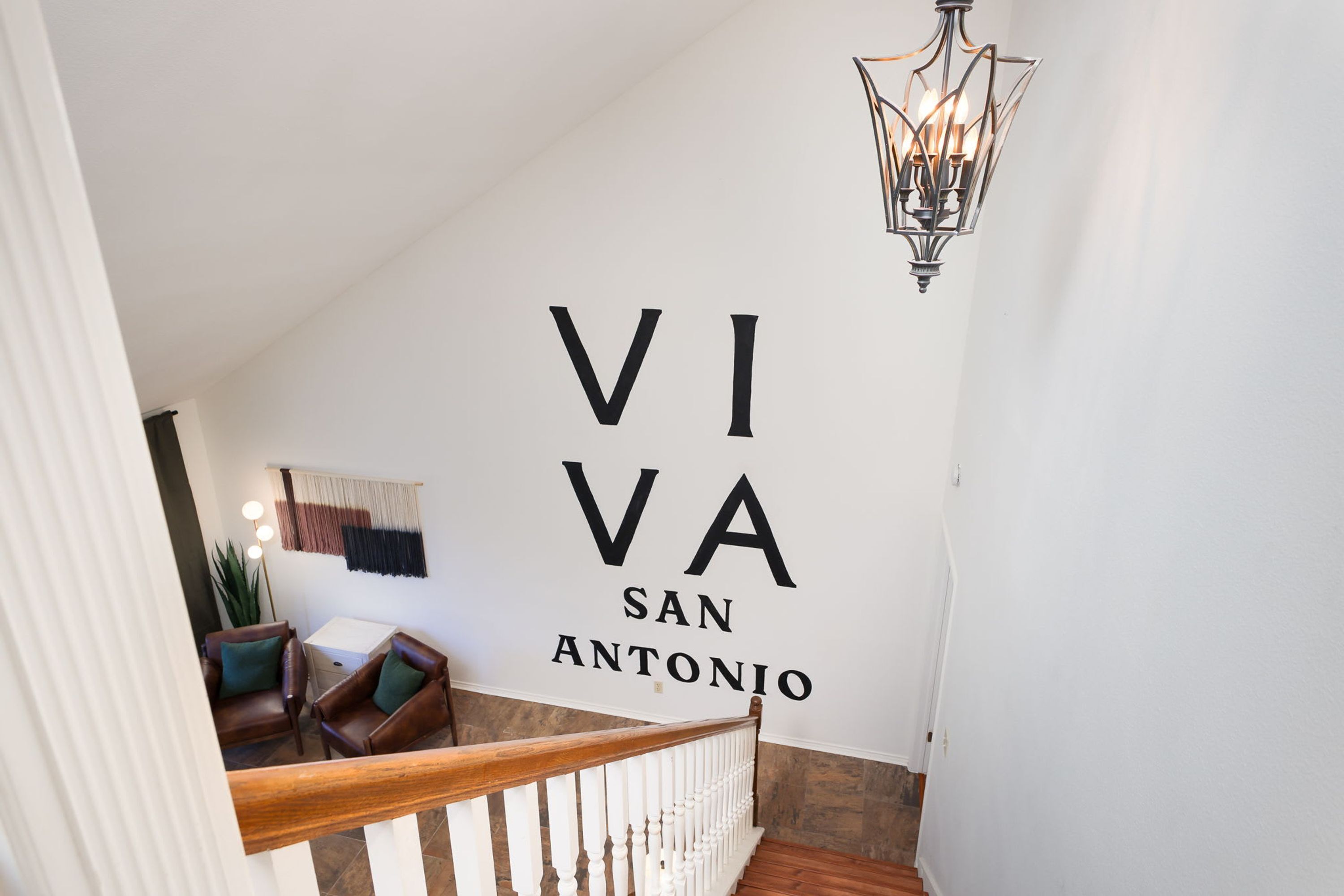 A staircase with a wooden handrail and white balusters, leading to a room with a large wall sign reading 'VIVA SAN ANTONIO'.