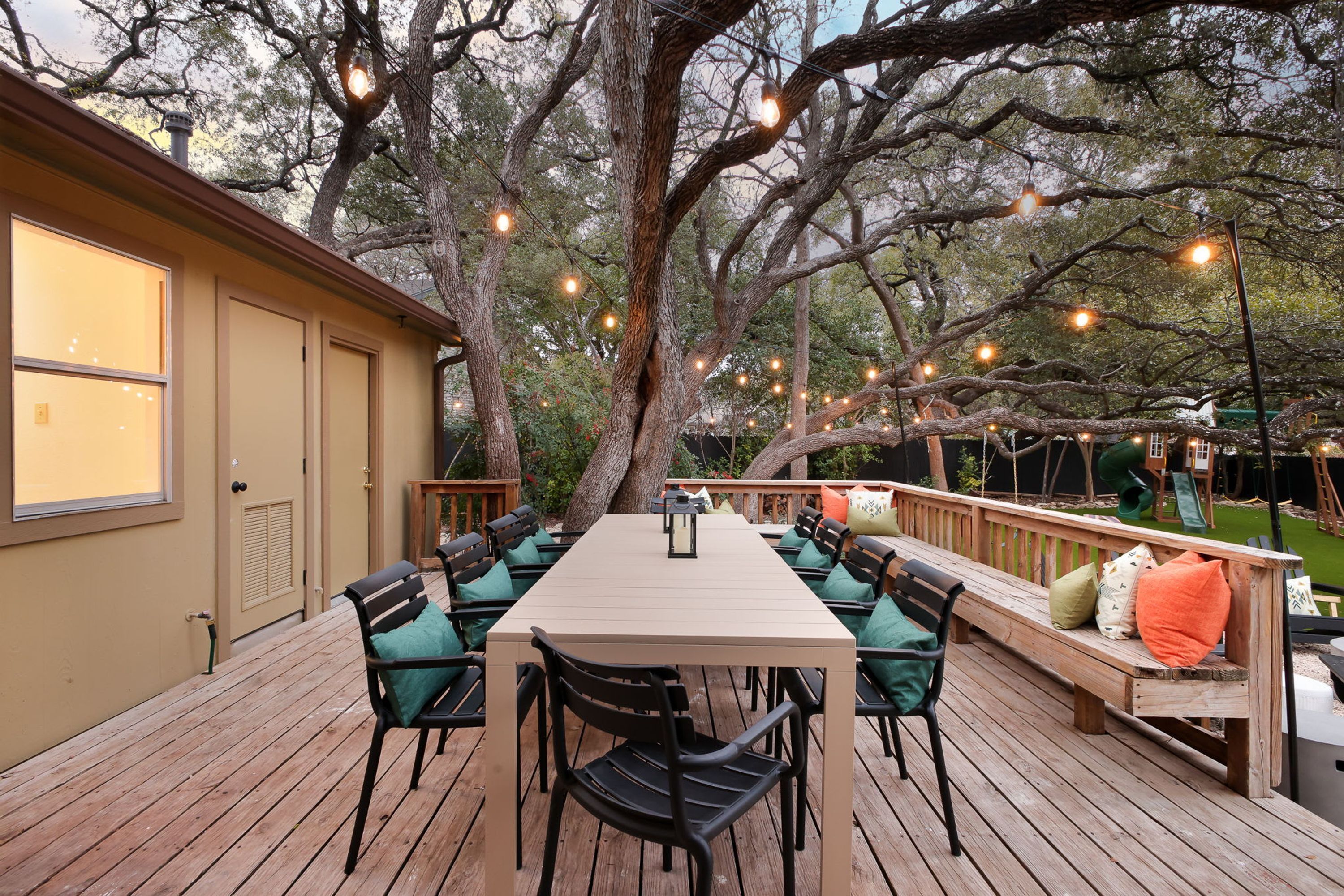 An outdoor deck with a long table and chairs, surrounded by trees and string lights.